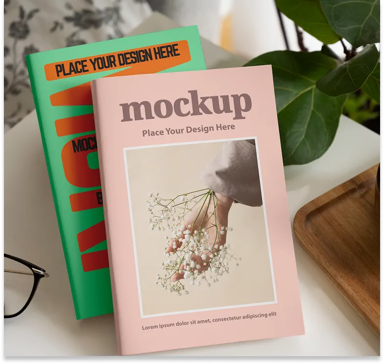 Two stacked magazines on a white table, the top one pink with a hand holding white flowers on the cover, the bottom one green with bold orange text.